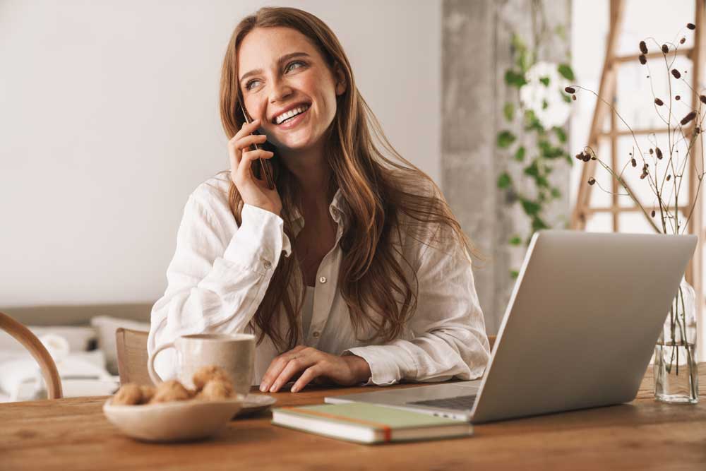 Das Foto zeigt eine lachende Frau die mit dem Handy telefoniert und vor einem Laptop sitzt unddient als Beitragsbild zum kostenlosen Umzugsratgeber 'Was ist vor einem Umzug zu beachten'.